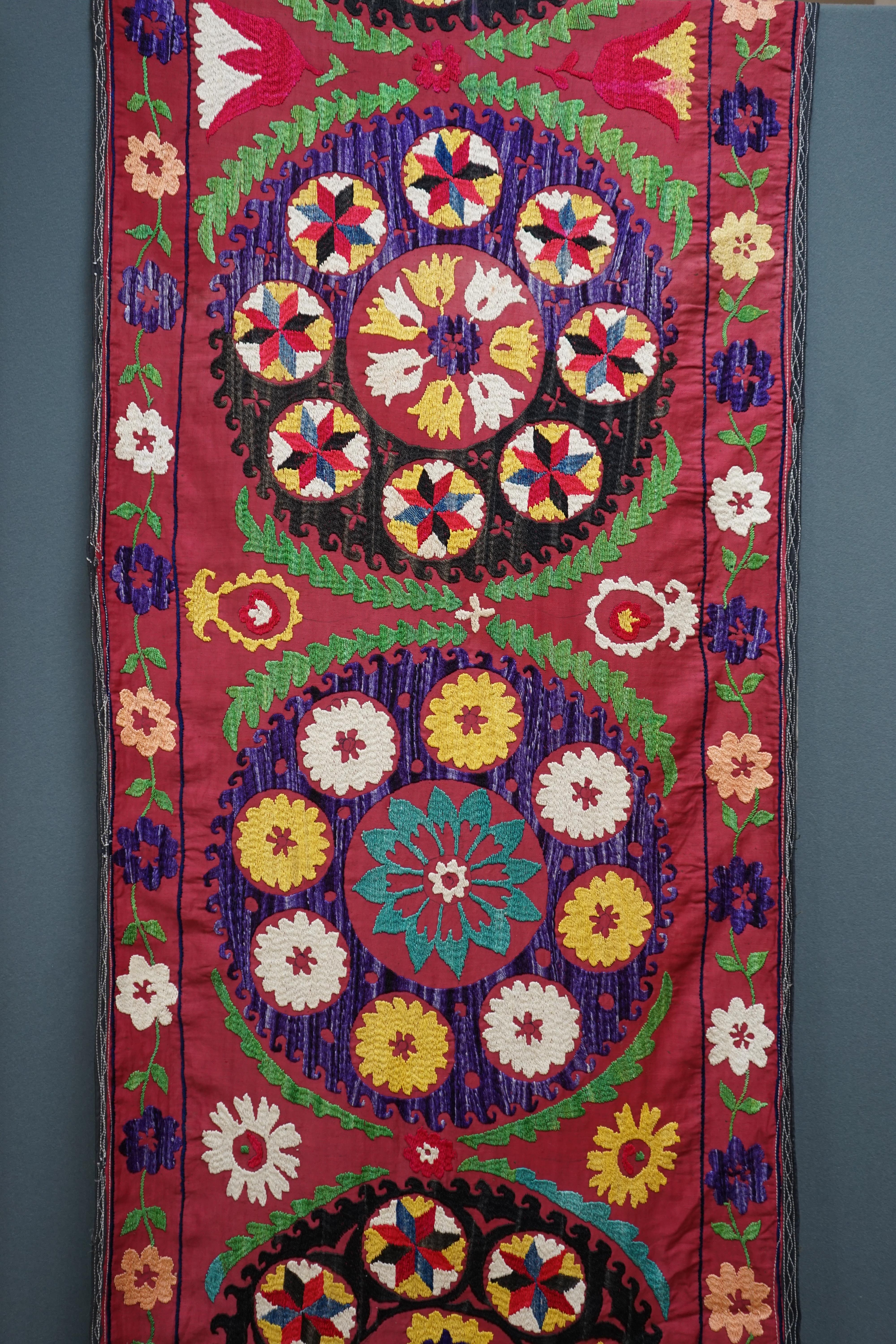 A 20th century Suzani wall hanging, possibly Uzbekistan, embroidered in bright polychrome silk threads into large symbols representing happiness, health and luck, on a burgundy cotton background, with a fine muslin backi